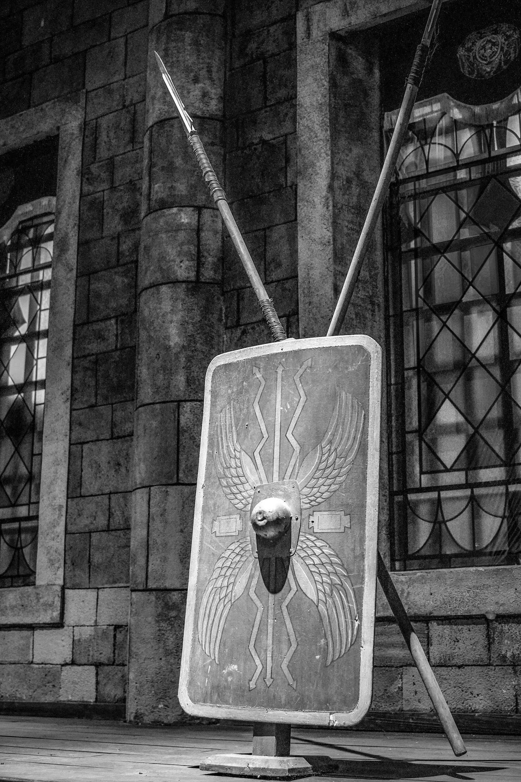 Black and white photograph of a Roman shield, symbolizing defense, and a spear leaning against it, set against a brick building with barred windows.