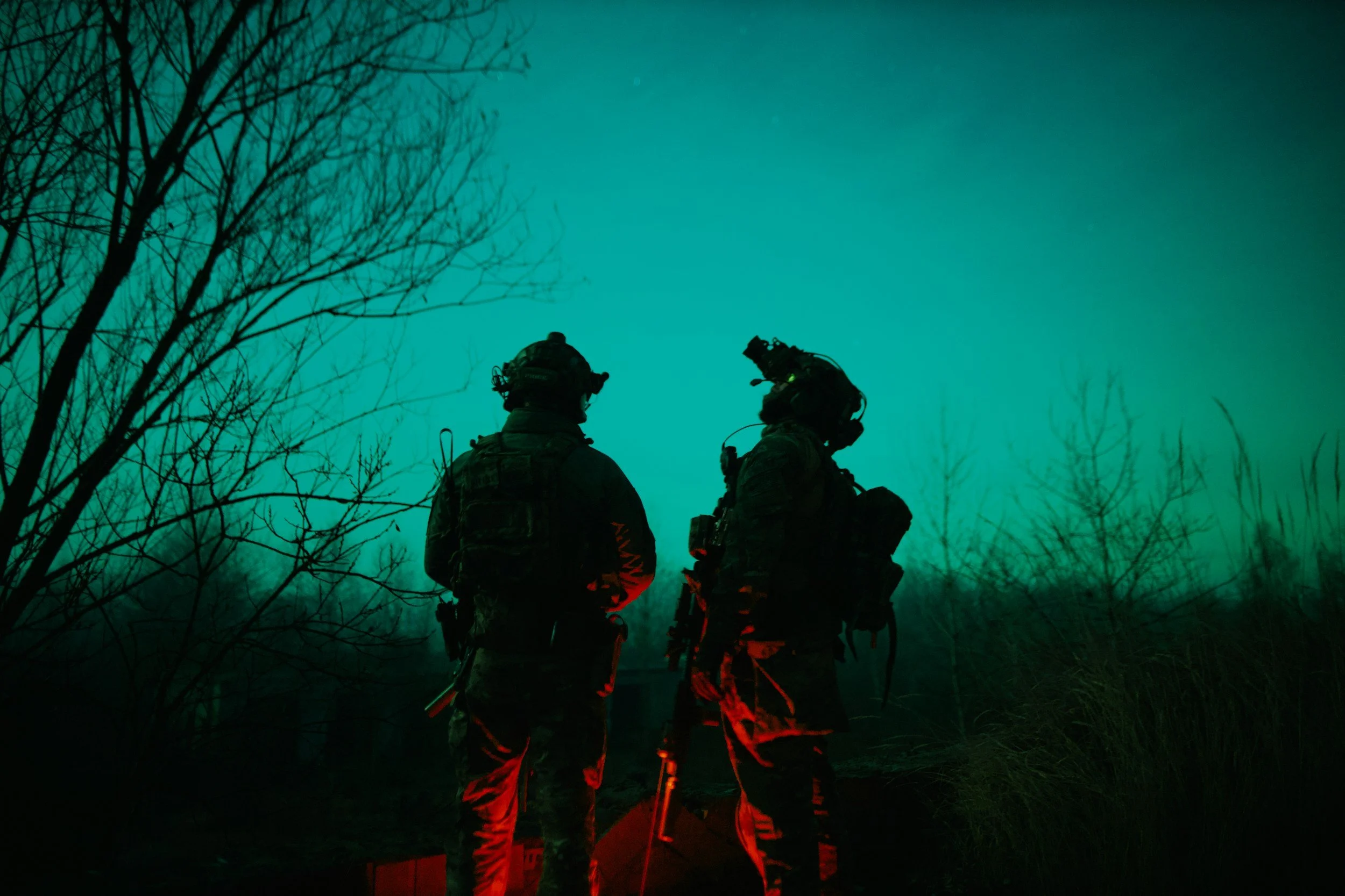 Two soldiers in tactical gear standing outdoors at night, illuminated by red and green lights, with leafless trees in the background.