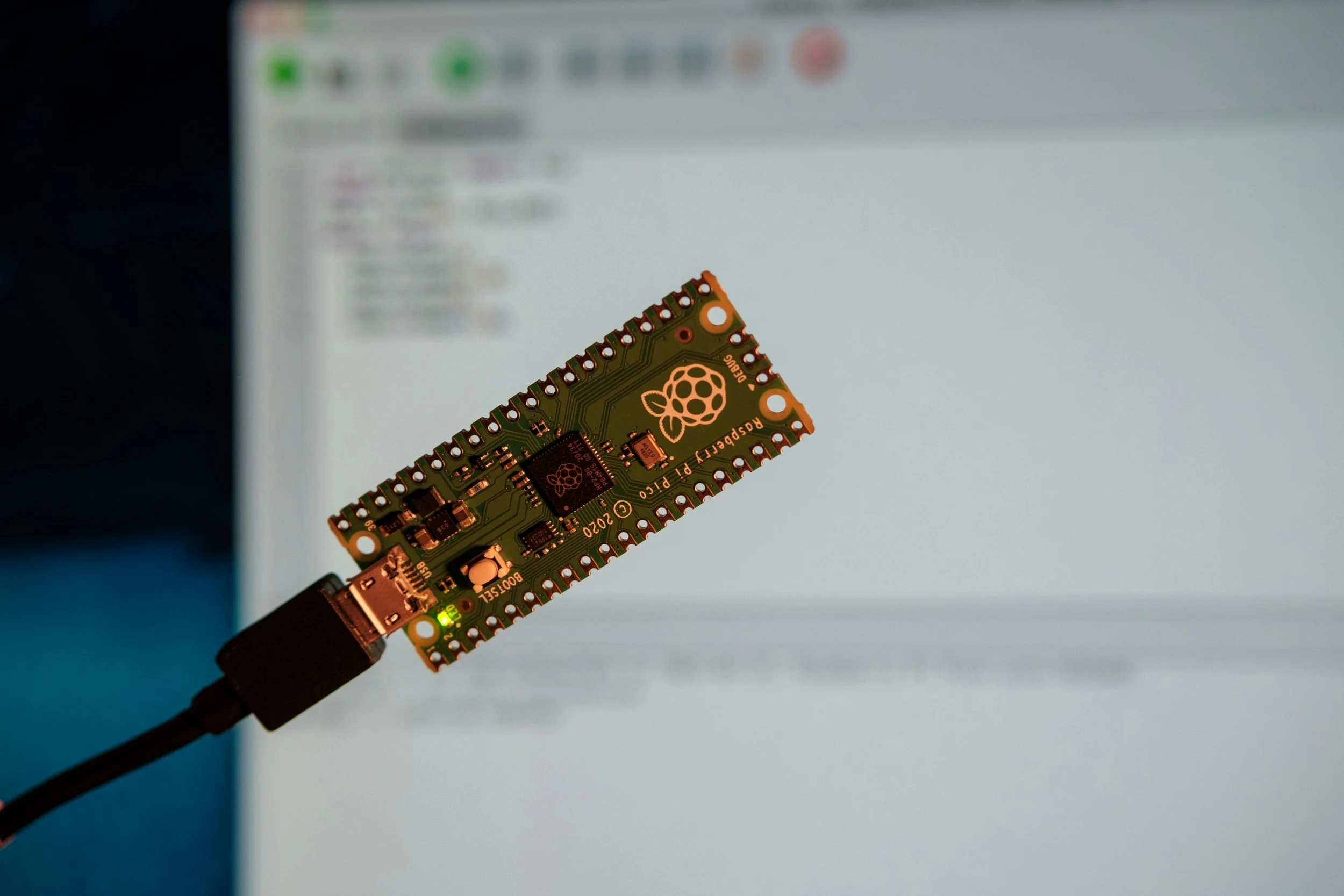 Close-up of a Raspberry Pi microcontroller with green circuit board and copper components, connected via a black cable in front of a blurred computer screen.