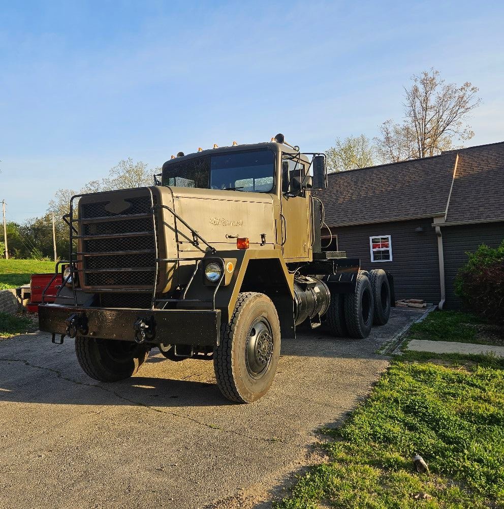 A large, vintage military truck parked in a driveway next to a house with gray siding and a small window. The truck is painted in an olive drab color and has a sturdy metal grille and bumper. Trees and a clear blue sky are visible in the background.