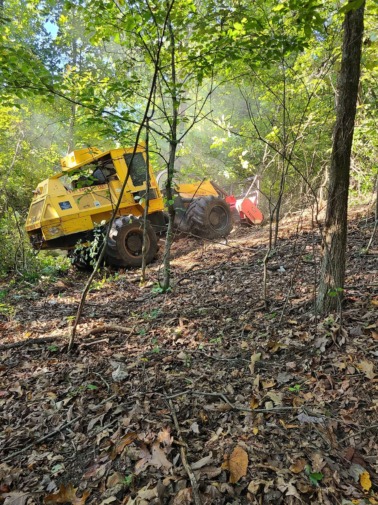 A yellow and red leaf blower operating in a wooded area with green leaves and trees, with a dusty trail behind.