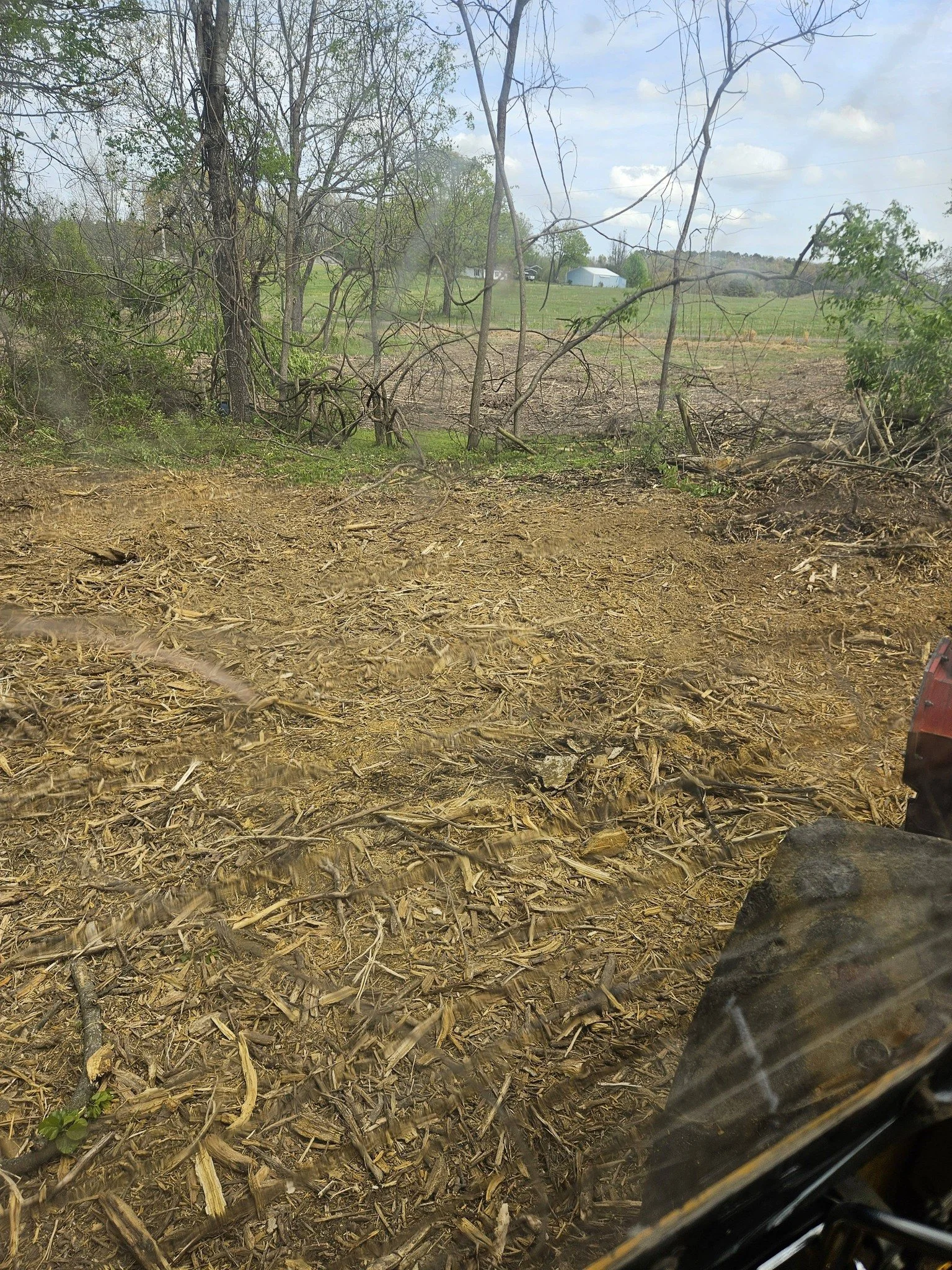 View from inside a vehicle showing a partially cleared area with wood debris on the ground, trees, open fields, and a metal building in the background under a partly cloudy sky.