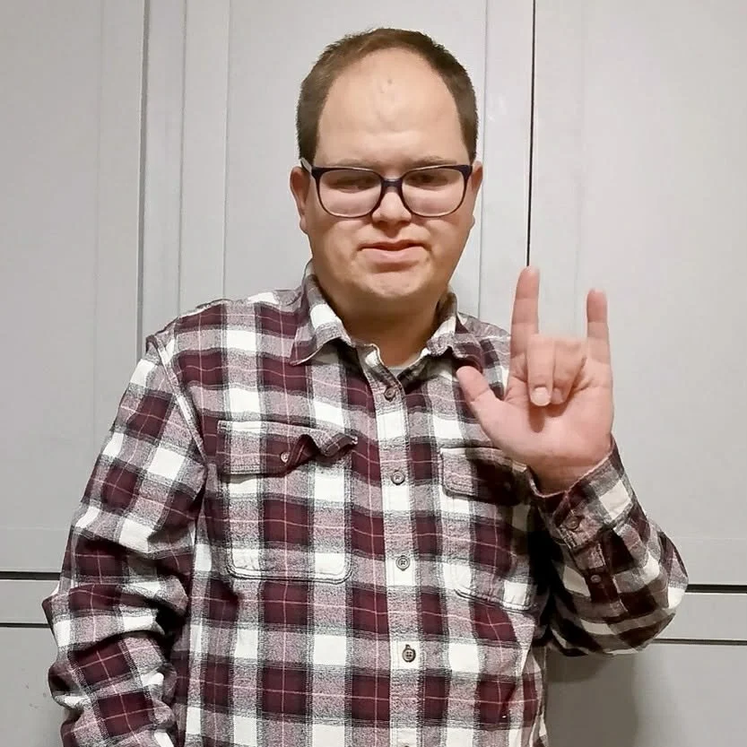 A man with glasses in a plaid shirt making a hand gesture with his right hand, standing in front of a plain light-colored wall.