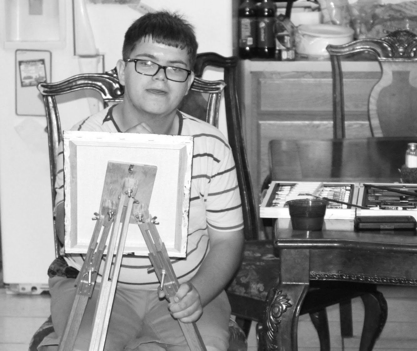 A young man with glasses and short hair sits at a dining table, holding a small easel with a blank canvas. The background includes a kitchen with various items on the counter, and a wooden table with art supplies, including a paint palette and contai