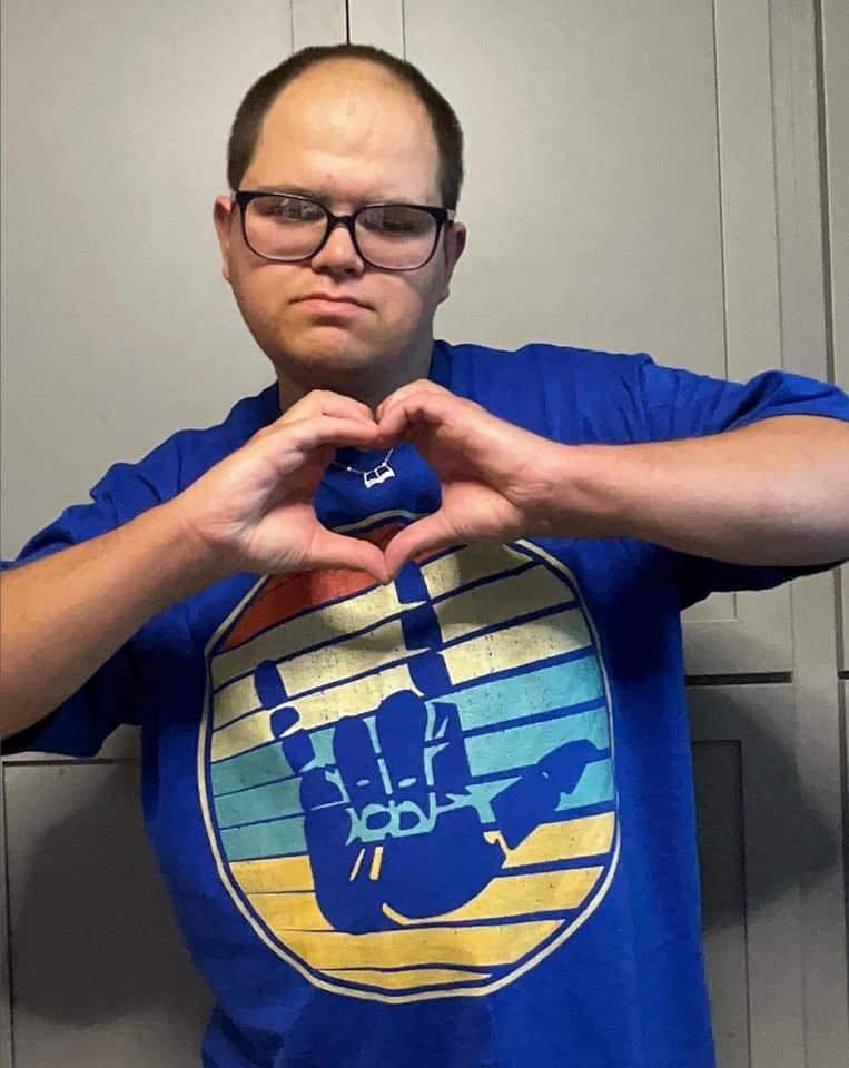 A man wearing glasses making a heart shape with his hands in front of his chest, standing against a gray background, wearing a blue T-shirt with a graphic of a hand making a rock and roll gesture inside a circle with a gradient background.