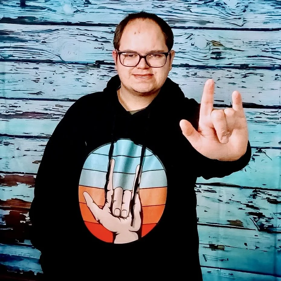 A person wearing glasses and a black hoodie with a colorful graphic of a hand making a rock and roll sign, standing in front of a wooden background. The person is holding up their right hand making the same rock and roll sign.