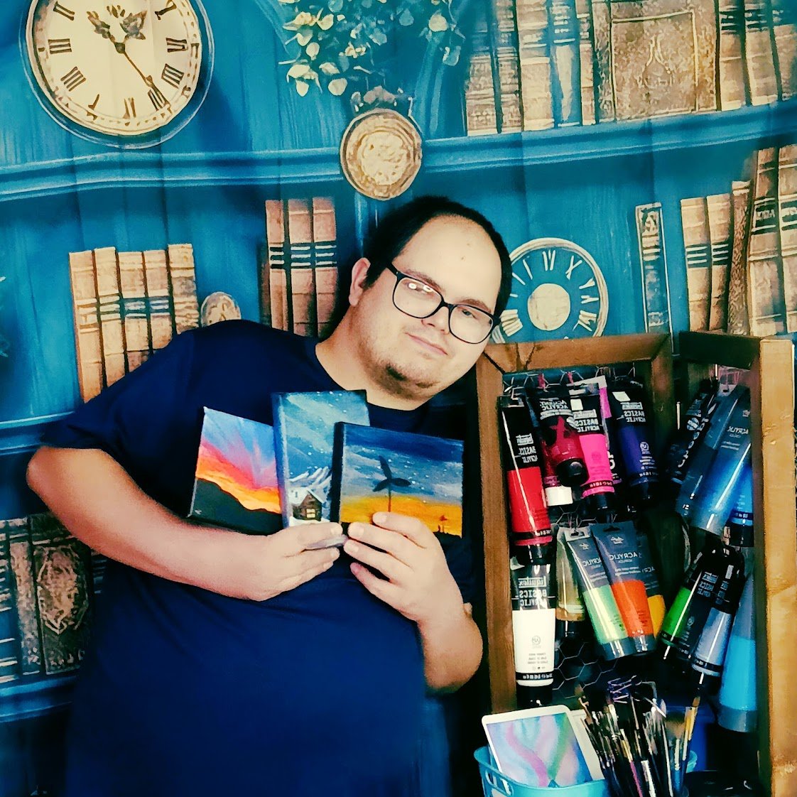 A person with glasses holding three landscape paintings, standing in front of a colorful, book-themed wall and a shelf with acrylic paint tubes.