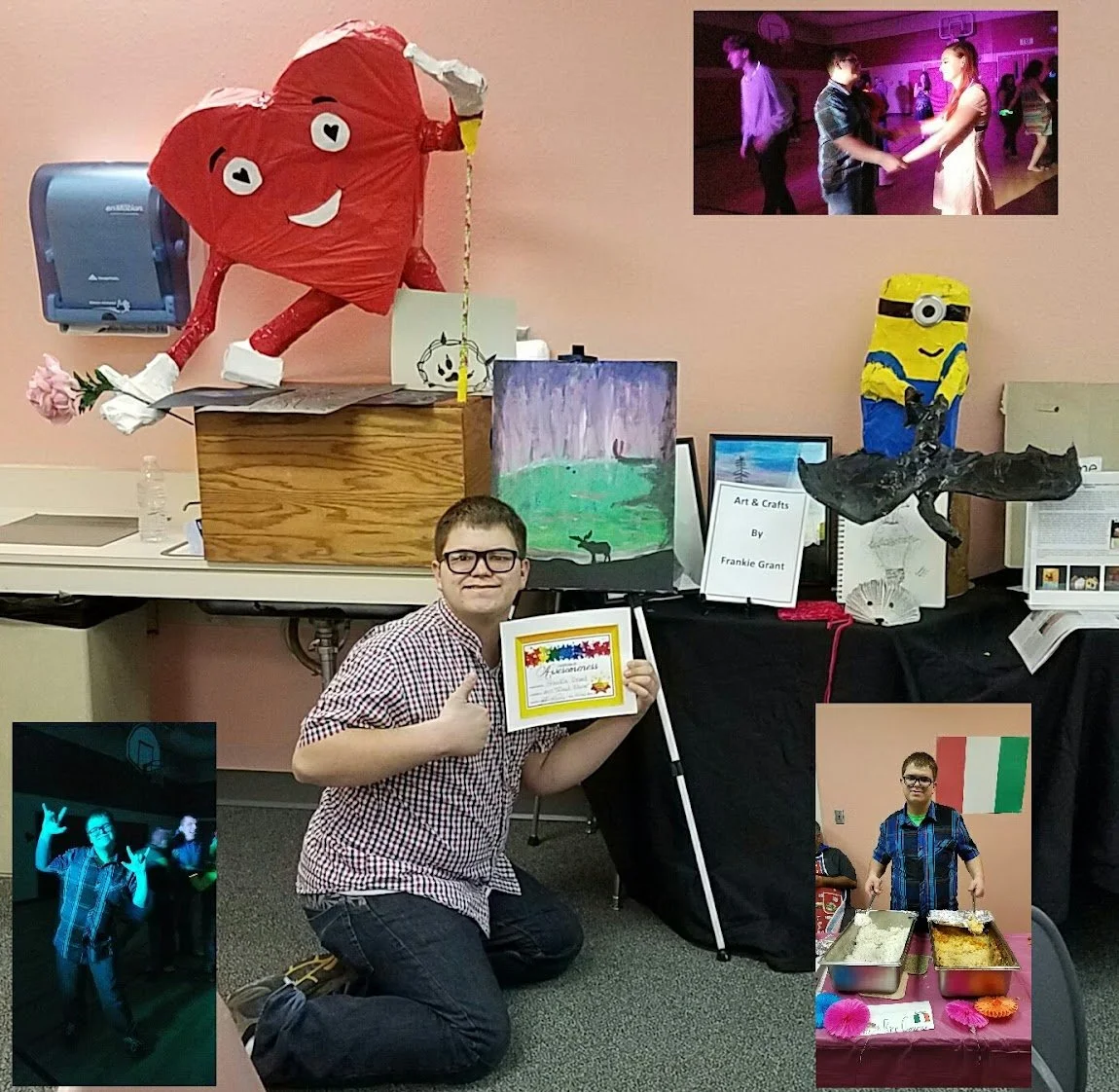 A young boy kneeling on the floor holding a framed certificate, giving a thumbs-up at a school arts and crafts display. The display includes a large red heart-shaped balloon with a face, a Minion piñata, a painting of a deer in a green field, and sev