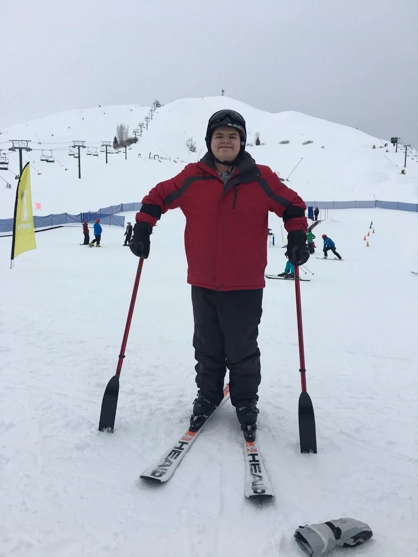 Person standing on skis with ski poles at a snowy ski resort, wearing a red jacket and black helmet, with ski lifts and other skiers in the background.