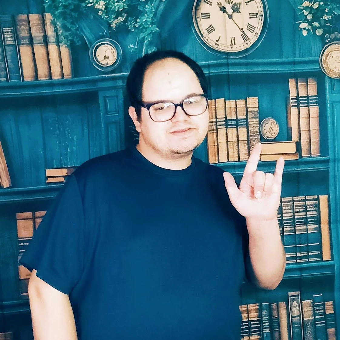 A man with glasses and a black t-shirt making a rock hand sign in front of a bookshelf painted on a wall.