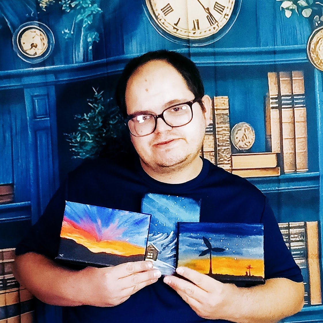 A man with glasses holding two small paintings of sunset scenes in front of a blue bookshelf and clock background.