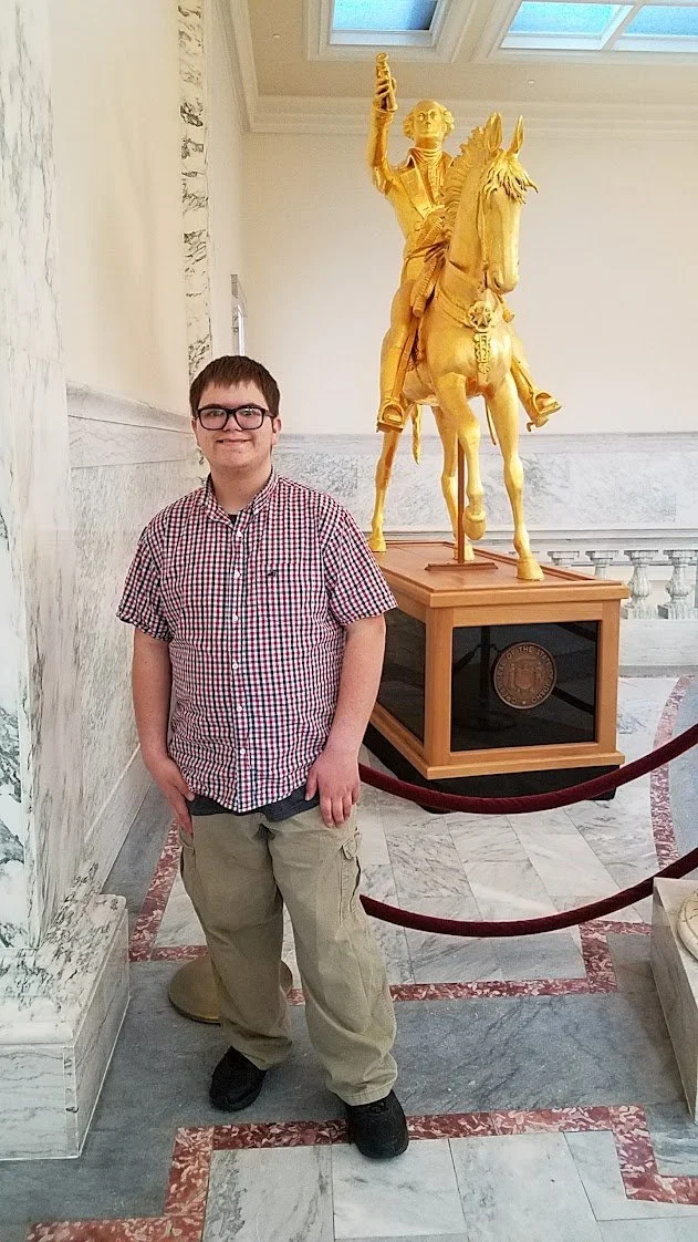 A young man wearing glasses, a red and white checkered shirt, and khaki pants standing inside a building with a marble floor, near a gold-colored sculpture of a man on a horse, holding a sword and a flag, in a historic or museum setting.