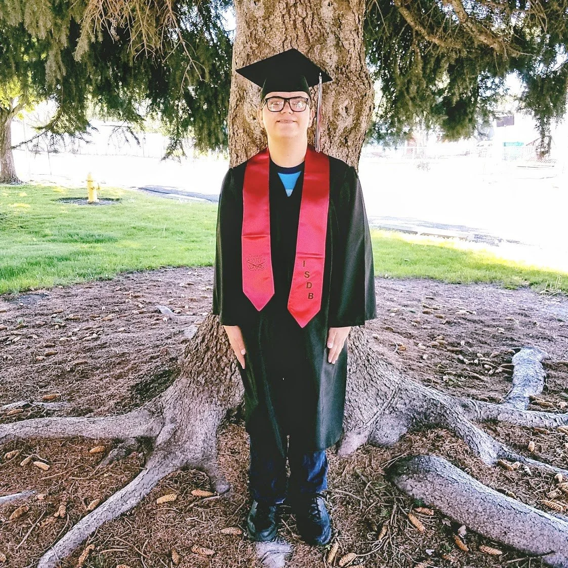 Young man in graduation cap and gown standing in front of a tree, holding an umbrella, with a red stole that reads "ISDB".