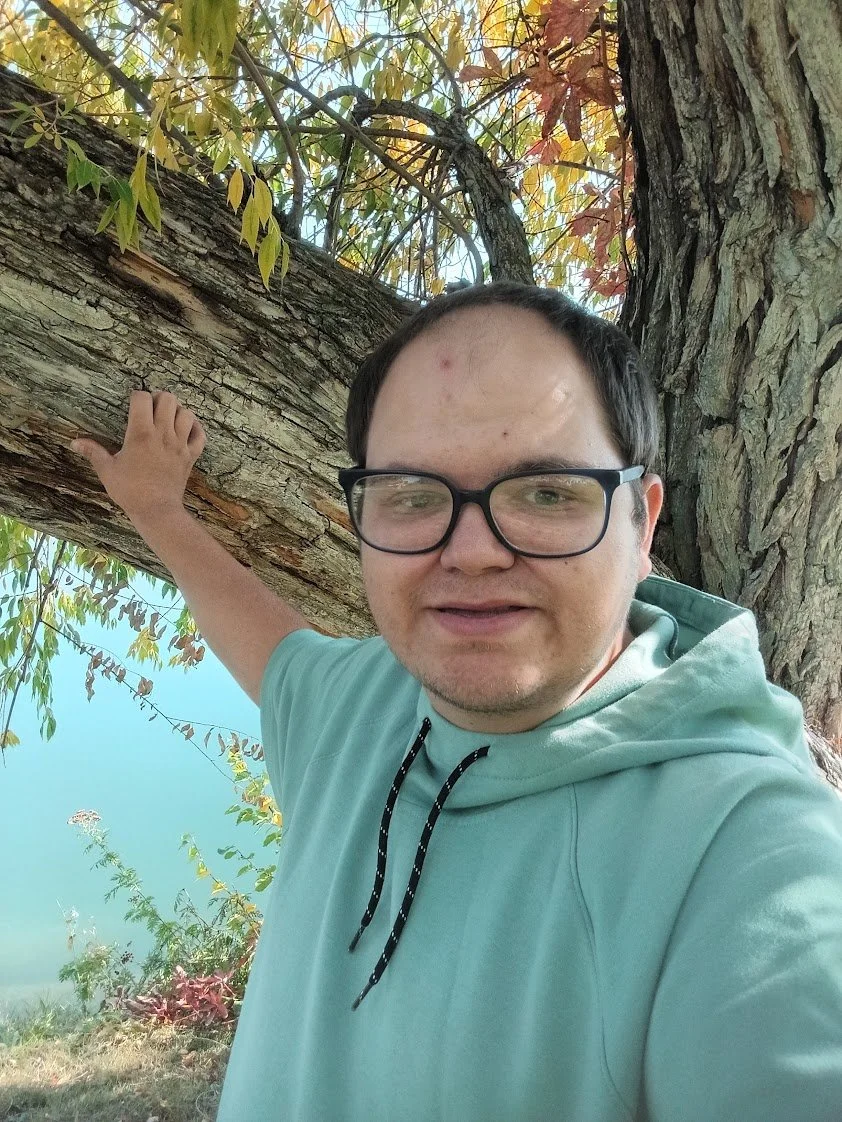 A man with glasses taking a selfie outdoors, hugging a large tree with his arm, with green and autumn leaves in the background.