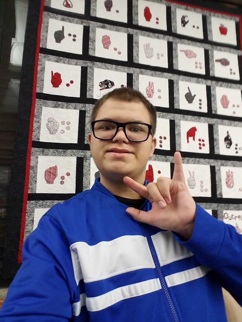 Young man wearing glasses and a blue and white jacket taking a selfie in front of a quilt display with various red, black, and white fabric patterns.