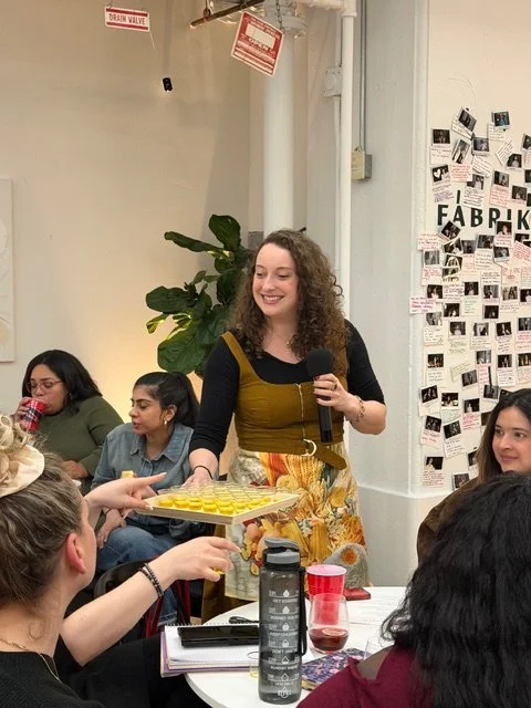 A woman with curly hair in a black and yellow dress, holding a microphone, reaching out to receive a yellow cupcake tray from a person in a black shirt, during a gathering in a room with a white wall and a collage of photos labeled 'FABR'
