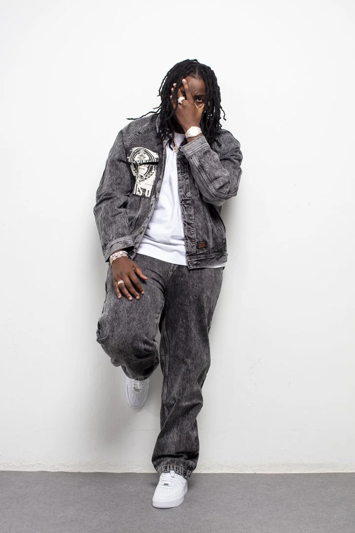 Person with dreadlocks wearing a gray denim jacket and matching gray pants, standing against a white wall, with one leg raised and covering part of their face with their hand.