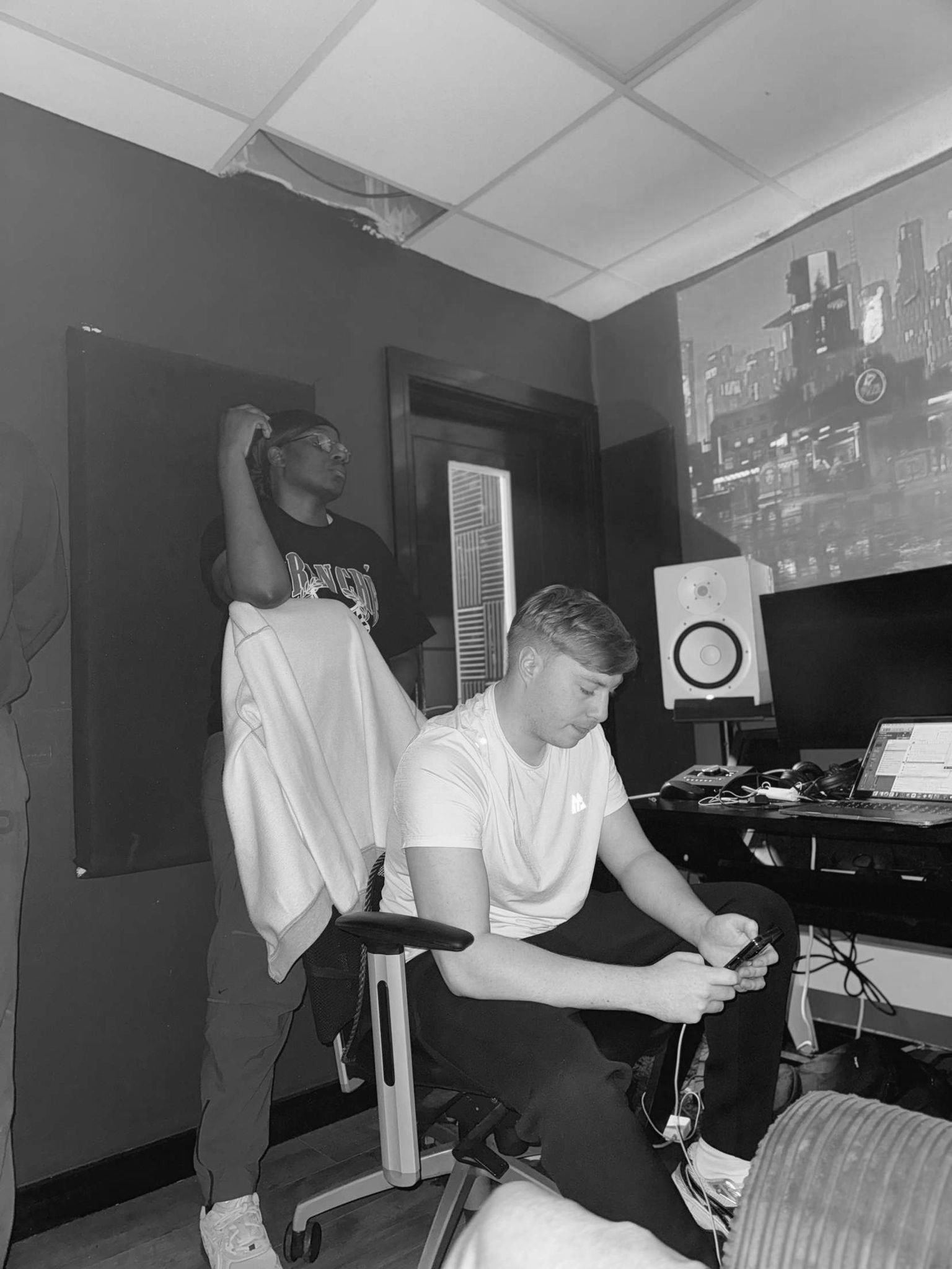 A person sitting in a chair using a smartphone, with another person standing behind, looking to the side, in a room with audio equipment and a projection of a cityscape on the wall.