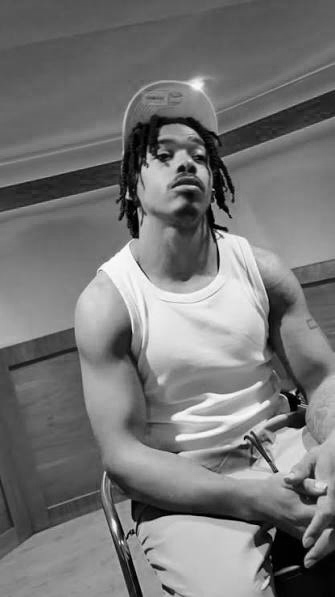 Young man with dreadlocks wearing a sleeveless shirt, seated indoors, looking at the camera.