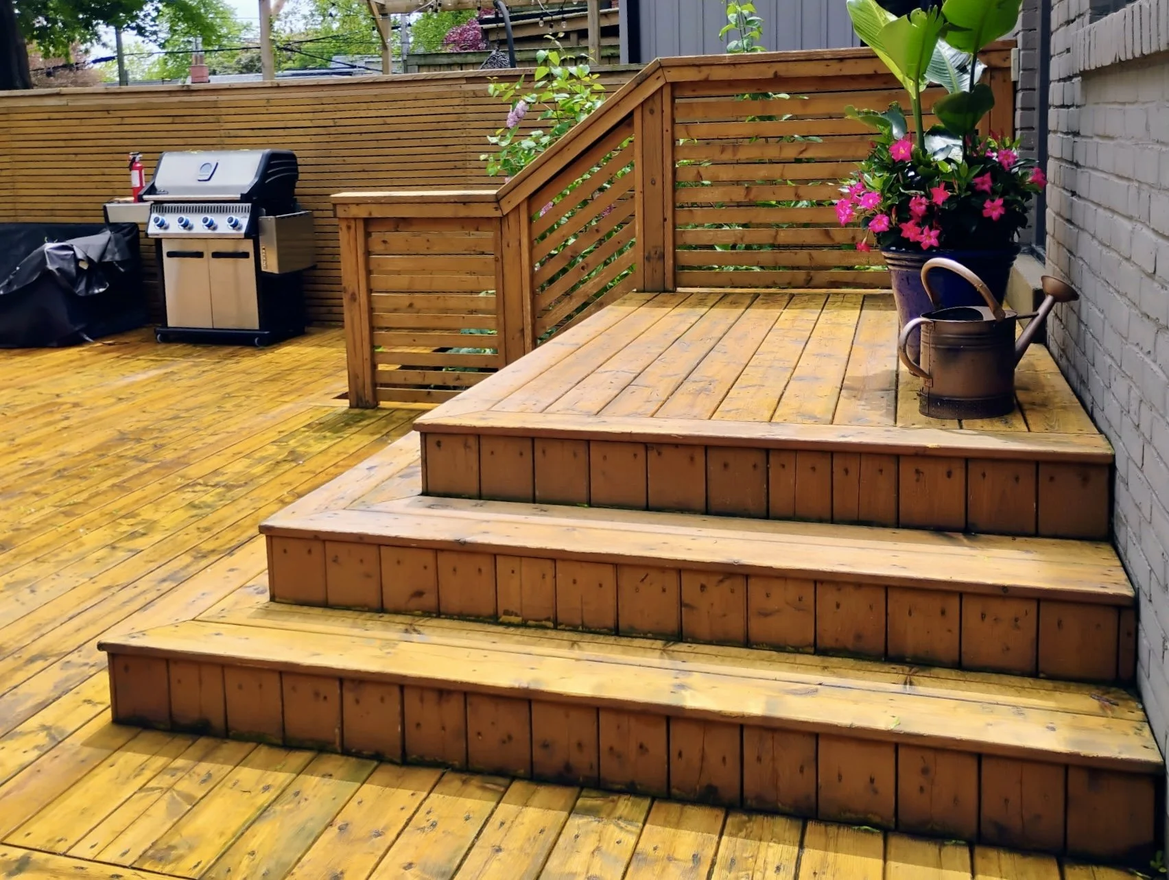 A wooden outdoor deck with stairs, surrounded by a wooden fence, a gray brick wall, and various potted plants, including a pink flowering plant, a watering can, and a barbecue grill, during daytime.