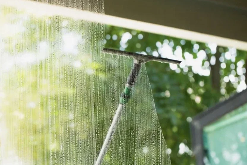 A window cleaning tool with a squeegee cleaning a glass window with water and soap, with droplets visible on the glass and trees outside in the background.