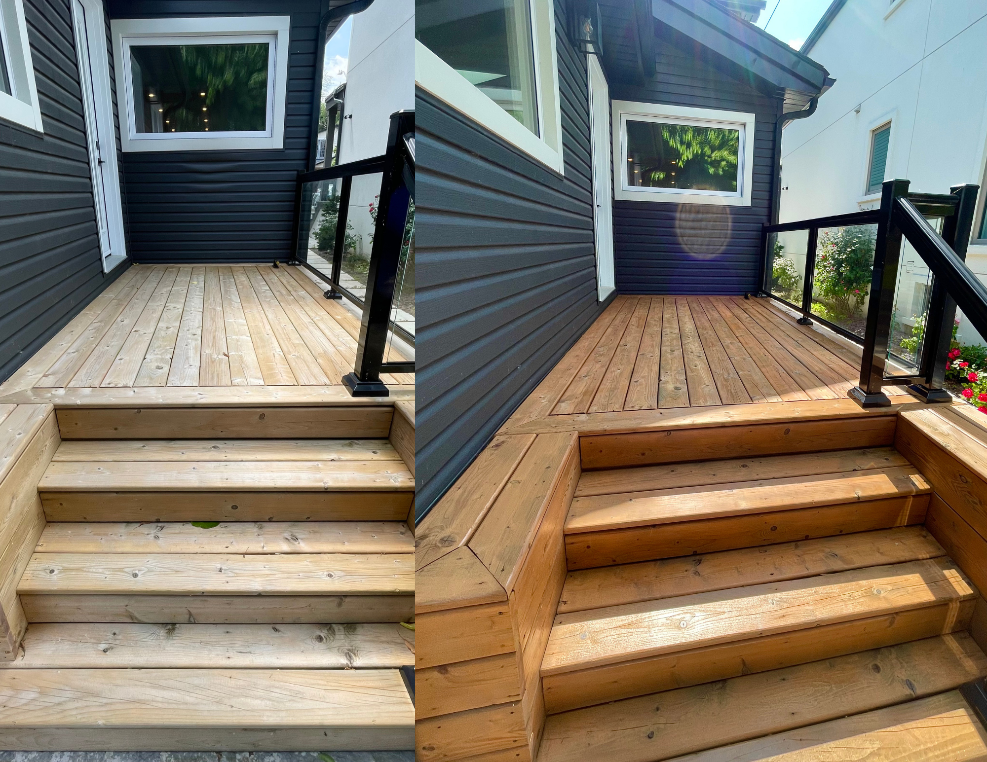 A small wooden deck with stairs leading up to it, attached to a house with dark gray siding and a white-framed window. The deck has a black metal railing with glass panels and is outdoors with plants and other houses visible in the background.