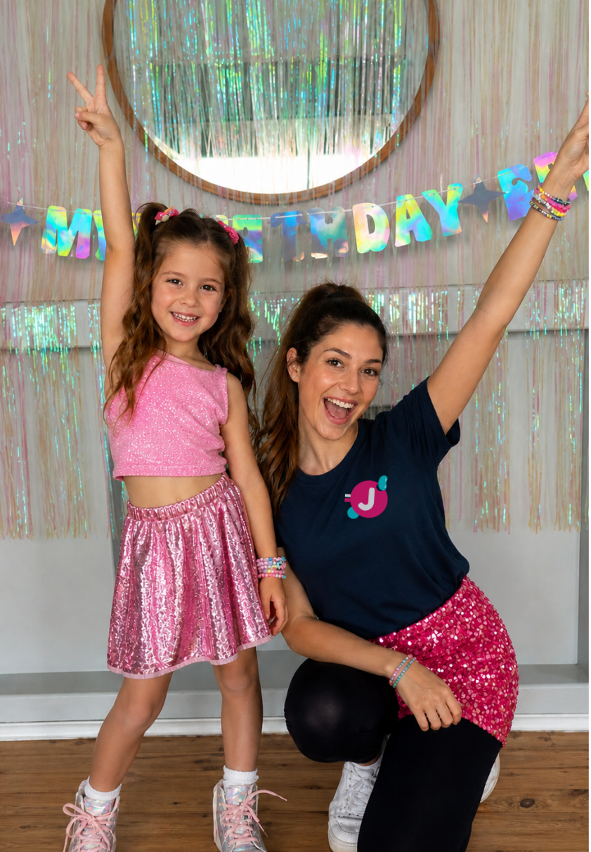 A young girl and woman celebrating a birthday indoors with a colorful 'Happy Birthday' banner and glittery decorations, both smiling and making peace signs.