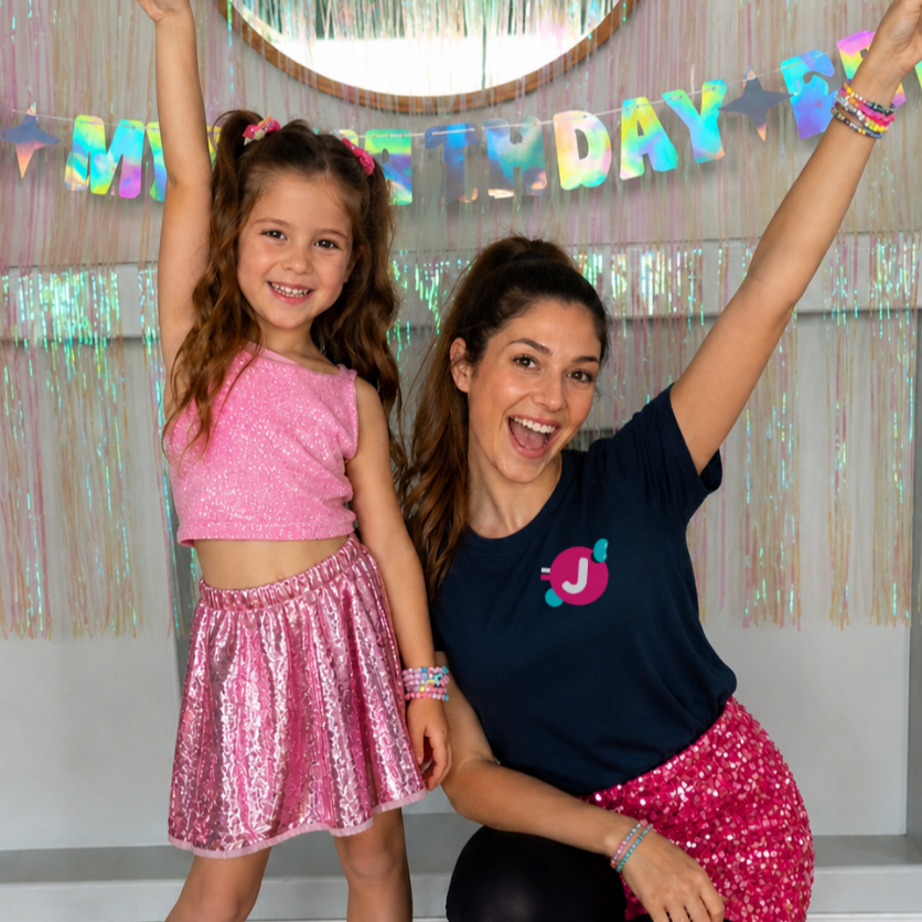 A woman and a young girl celebrating a birthday indoors, with colorful decorations and a 'Happy Birthday' banner in the background. The girl is wearing a pink sleeveless top and a shiny pink skirt, while the woman is wearing a navy blue shirt with a pink 'J' badge and a pink sequined skirt. Both are smiling and raising their arms in excitement.