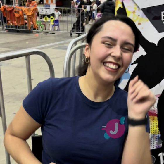 A woman with dark hair in a ponytail, wearing a blue t-shirt and gold hoop earrings, smiling and sitting against a metal barrier at an indoor event.