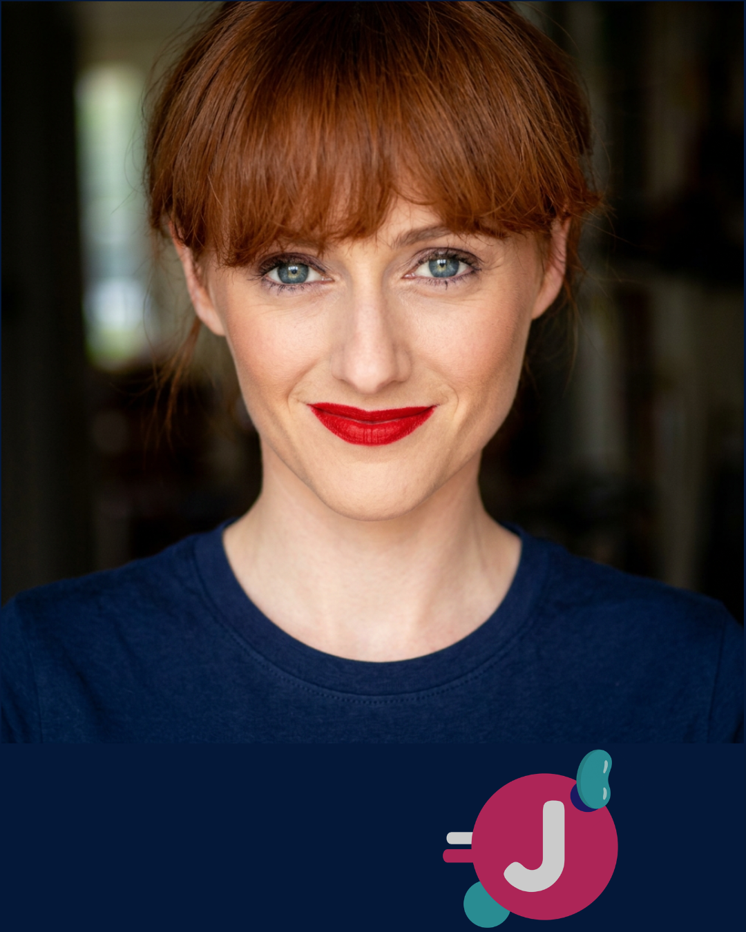 Close-up photo of a woman with red hair, blue eyes, and red lipstick smiling, wearing a navy blue shirt, with a blurred background.