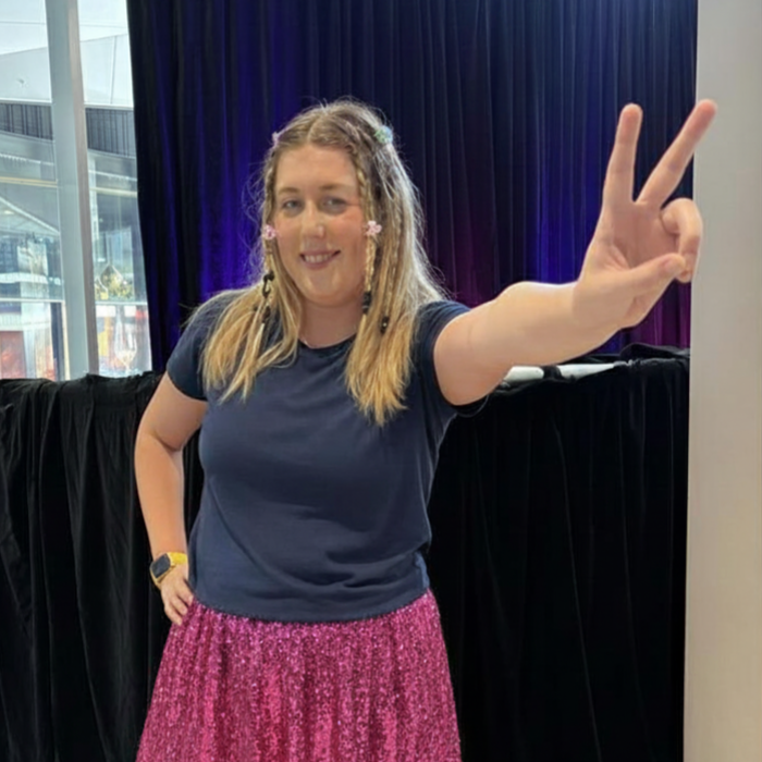 A young woman with long hair decorated with small flowers, wearing a navy T-shirt and a sparkly pink skirt, making a peace sign with her right hand and smiling at the camera indoors.