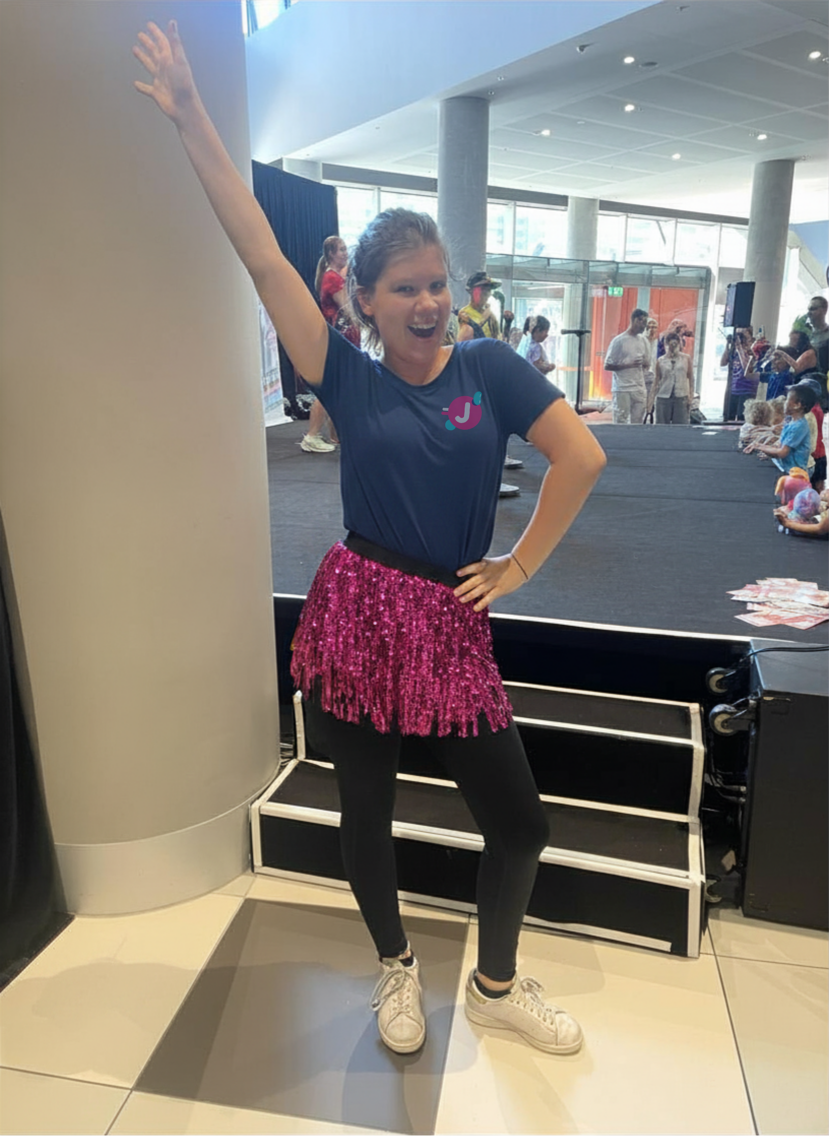 A young woman wearing a navy blue t-shirt, a pink sequined skirt, black leggings, and white sneakers posing with one arm raised and a big smile in a modern indoor setting with large windows and other people in the background.