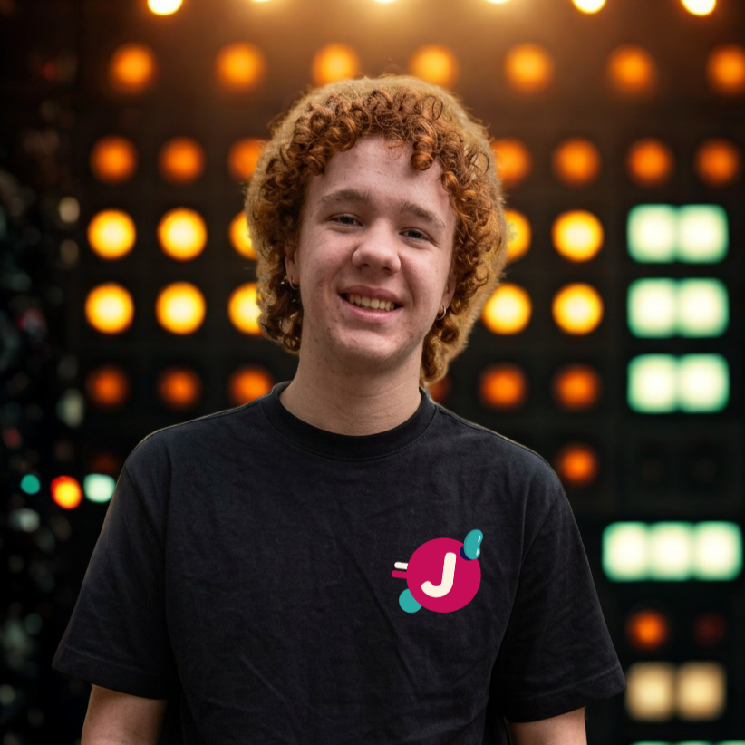 A young man with curly red hair, wearing a beige hat and a black t-shirt with a colorful logo on it, stands in front of a background of illuminated lights.