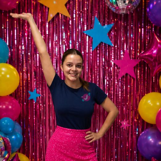 A young woman smiling and posing with one arm raised in front of a decorated party backdrop with colorful balloons, stars, and pink shiny curtains.
