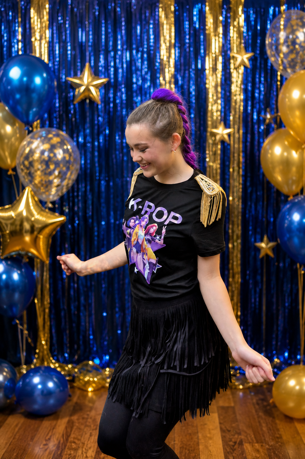 Young woman dancing with purple hair tied up, wearing a black K-pop t-shirt with animated characters and gold fringed epaulets, in front of a backdrop of blue and gold balloons and shiny blue and gold curtains.