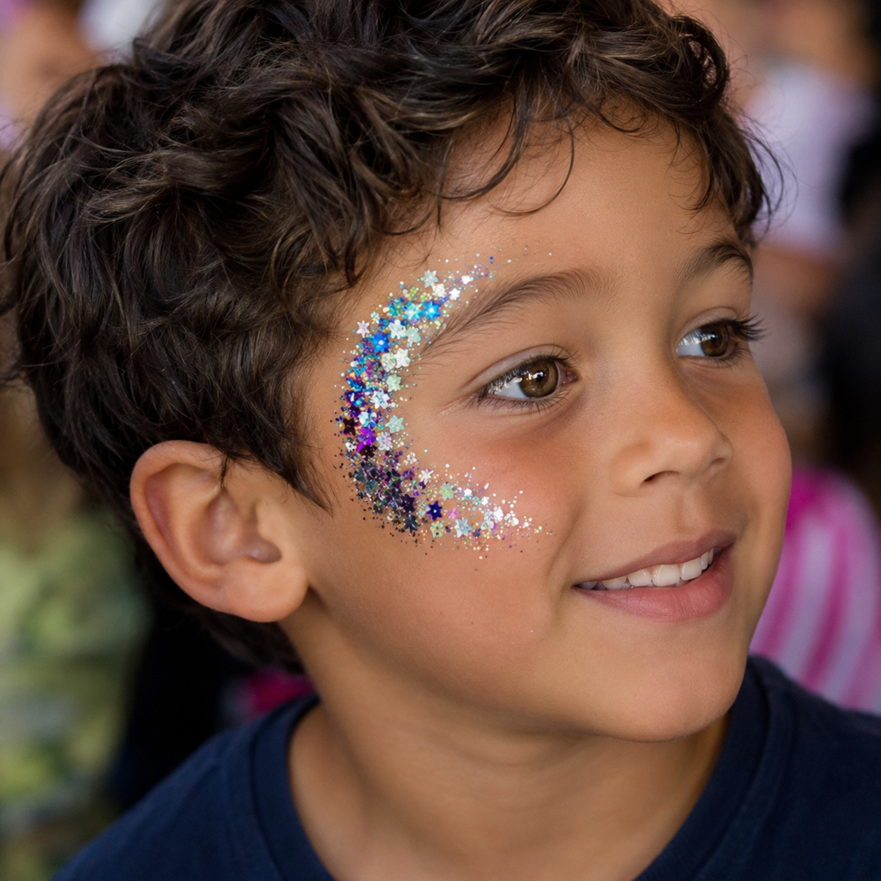 A young boy with curly brown hair and brown eyes has colorful glitter face paint along his cheek and eye area, forming a crescent shape. The background is blurred, suggesting an outdoor or crowded setting.