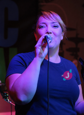 A woman singing into a microphone on stage, wearing a dark blue T-shirt with a small logo on the chest, illuminated by colored stage lights.