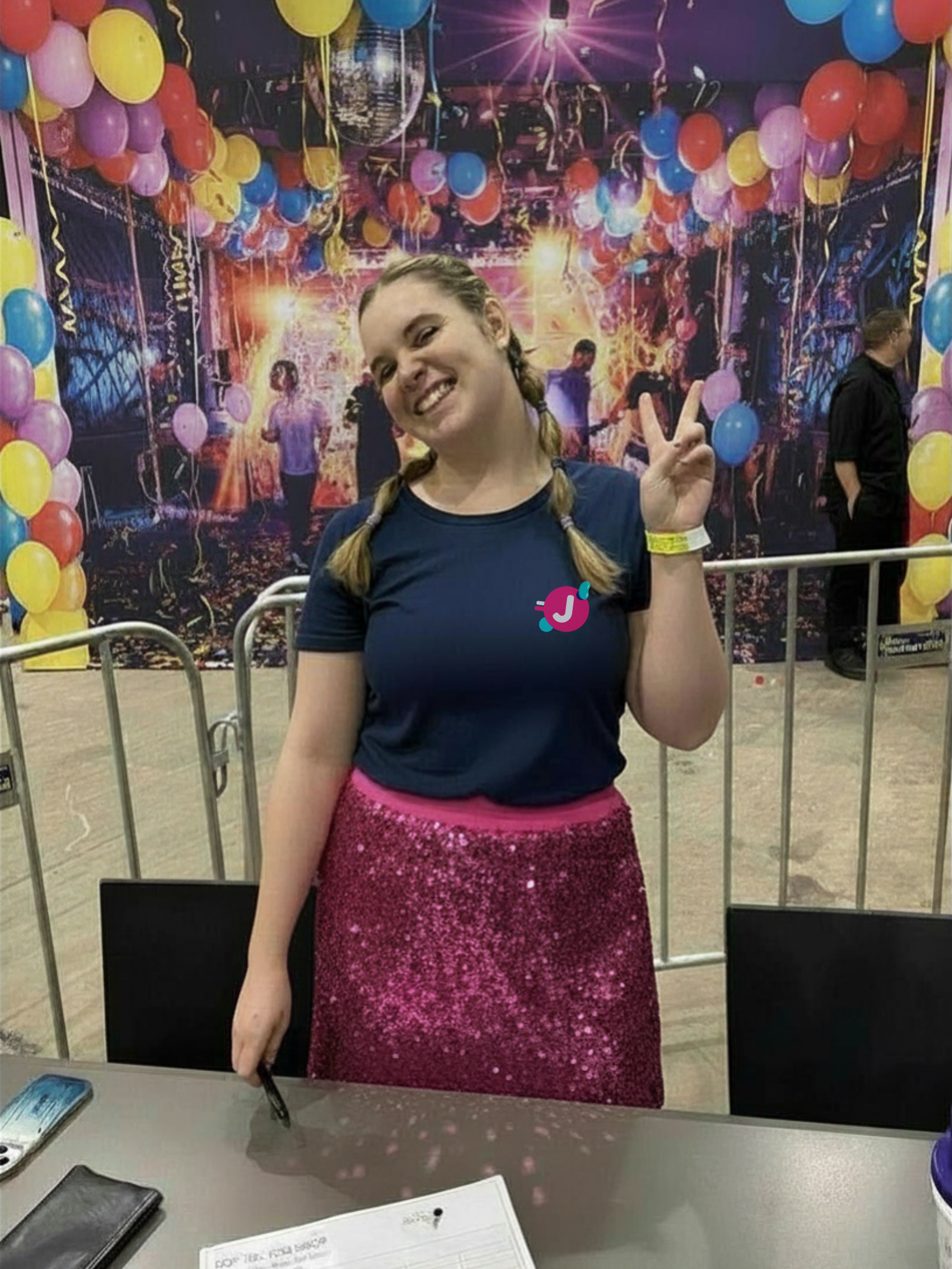 A young woman with braided hair is smiling and making a peace sign with her right hand. She is wearing a navy T-shirt and a glittery pink skirt. The background features a colorful party scene with balloons and a disco ball.