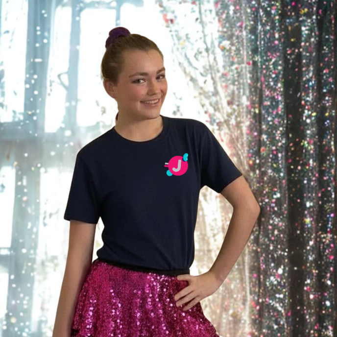 A young girl with a bun hairstyle wearing a navy t-shirt with a pink and blue logo and a shiny pink skirt, standing in front of a glittery curtain backdrop.