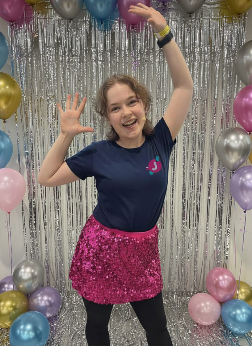 A young woman with short brown hair is smiling and waving at the camera, standing in front of a shiny silver tinsel backdrop decorated with balloons in pastel colors, including pink, blue, purple, silver, and gold, and wearing a navy blue T-shirt with a pink and blue logo and a pink sequined skirt.