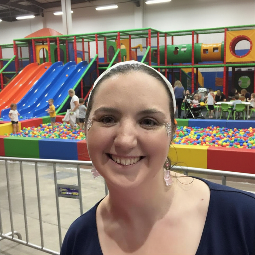 Smiling woman with glitter face paint and earrings, indoor playground with slides, ball pit, and children in the background.