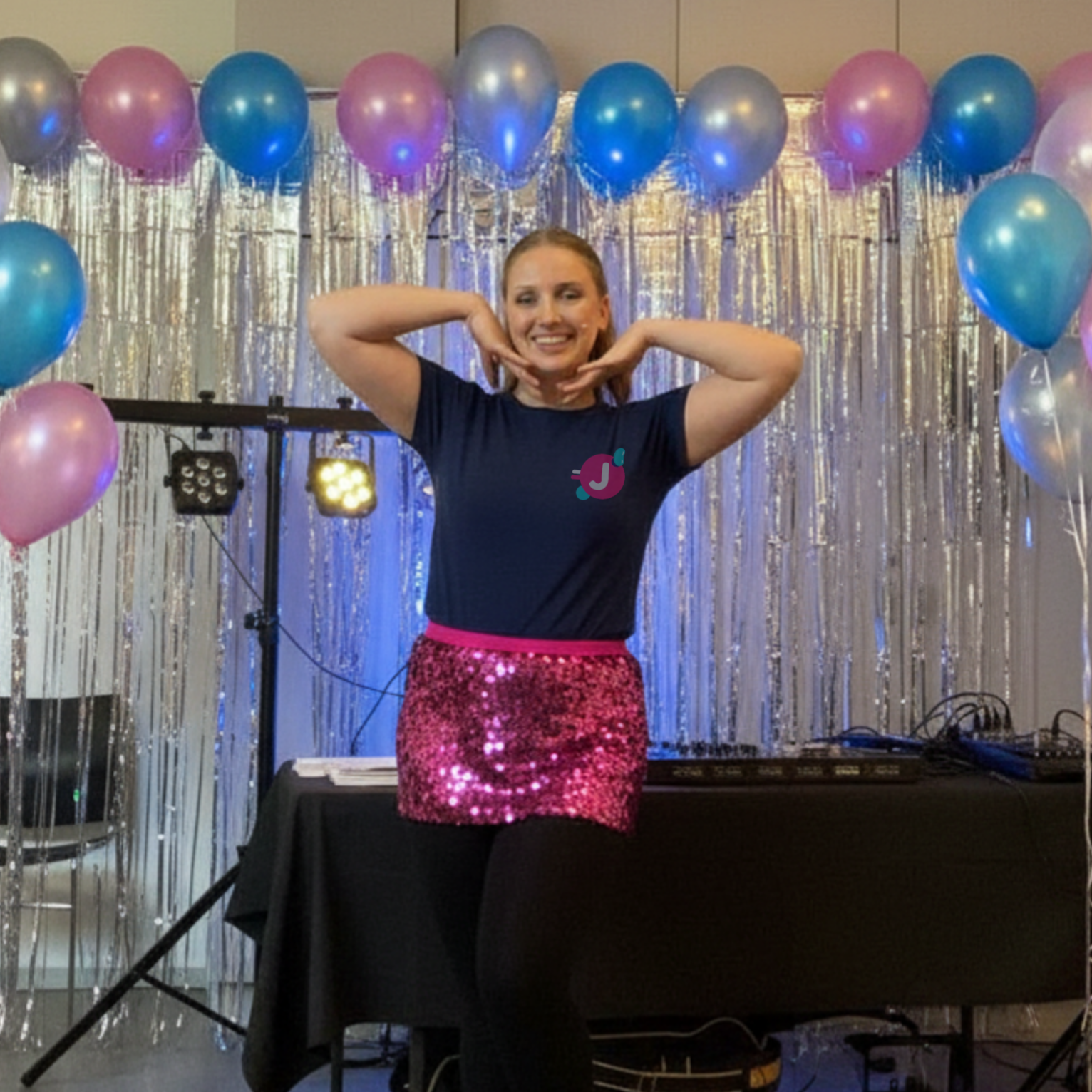 A woman posing with her hands under her chin in front of a backdrop decorated with balloons and streamers at an event or party.