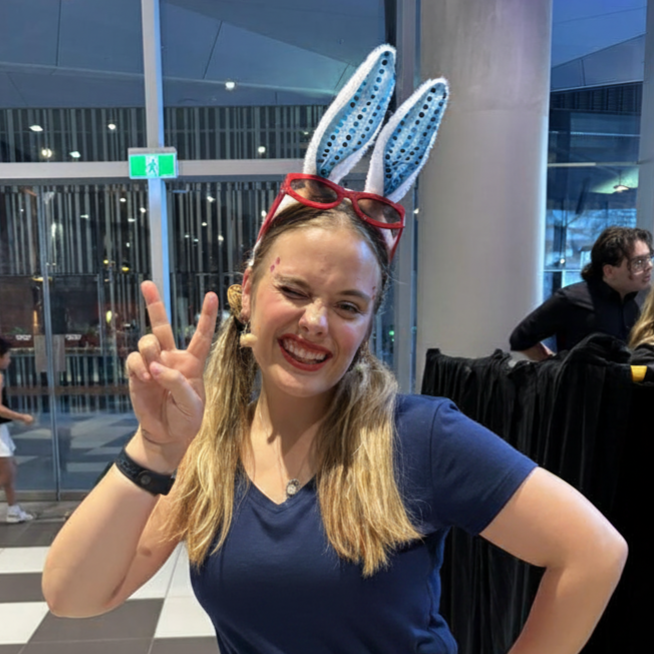 A young woman with long blonde hair wearing a blue T-shirt, red glasses, and bunny ears headband, making a peace sign and smiling at the camera.