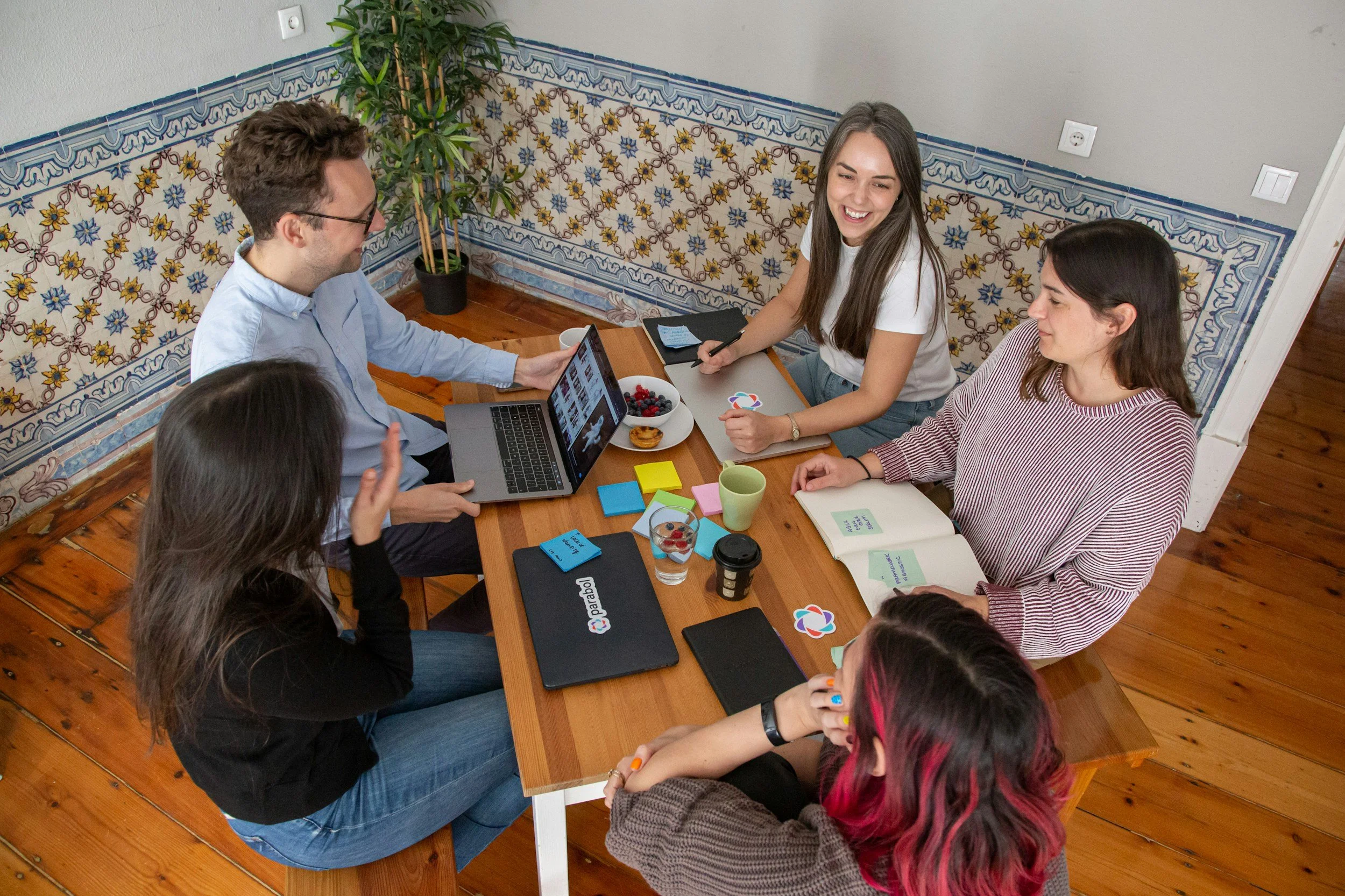 Group of six people sitting around a wooden table, engaging in a discussion with one person using a laptop, in a cozy room with patterned tiles on the wall and a potted plant.