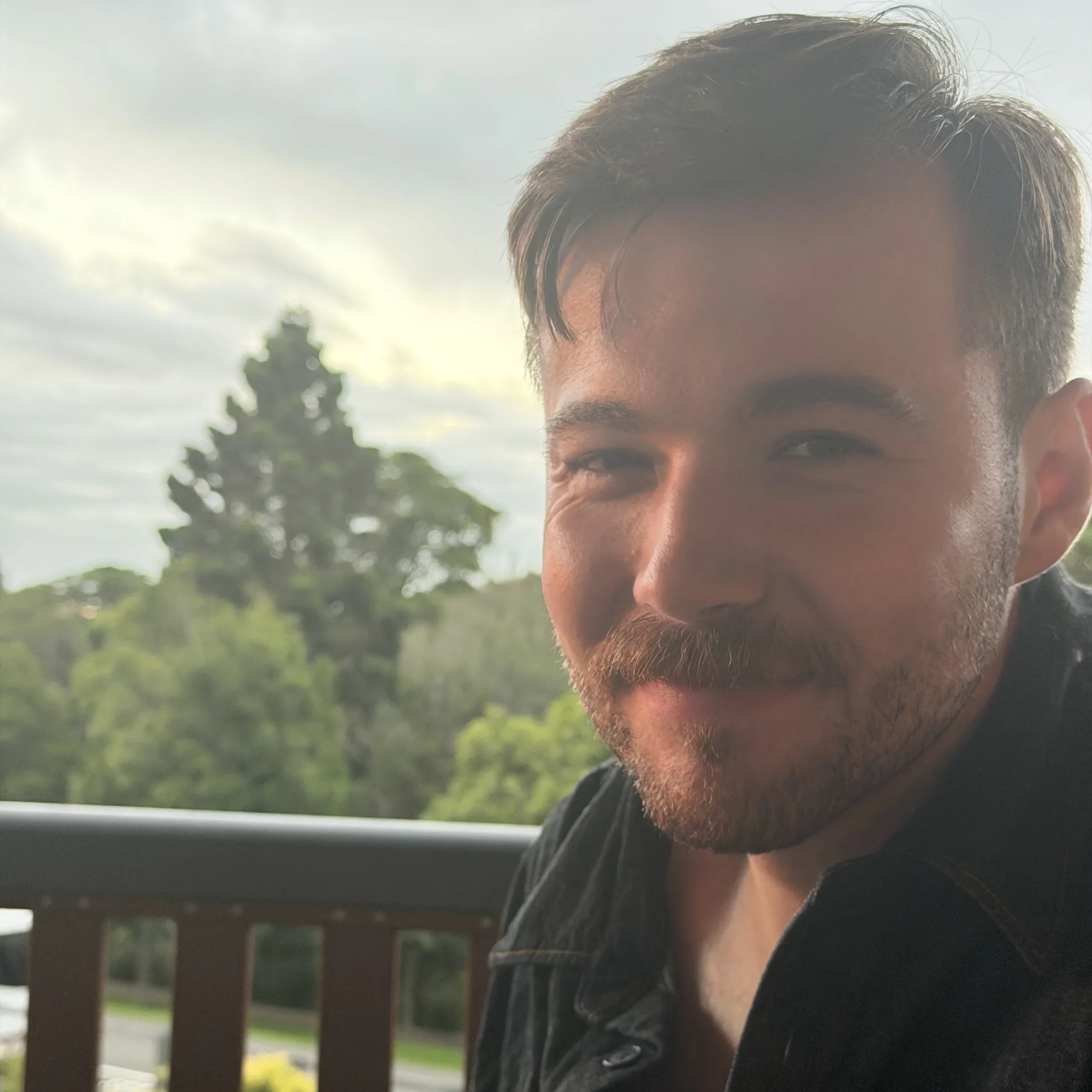 Close-up of a smiling man with short dark hair and a beard, standing outdoors near a porch with trees and a cloudy sky in the background.