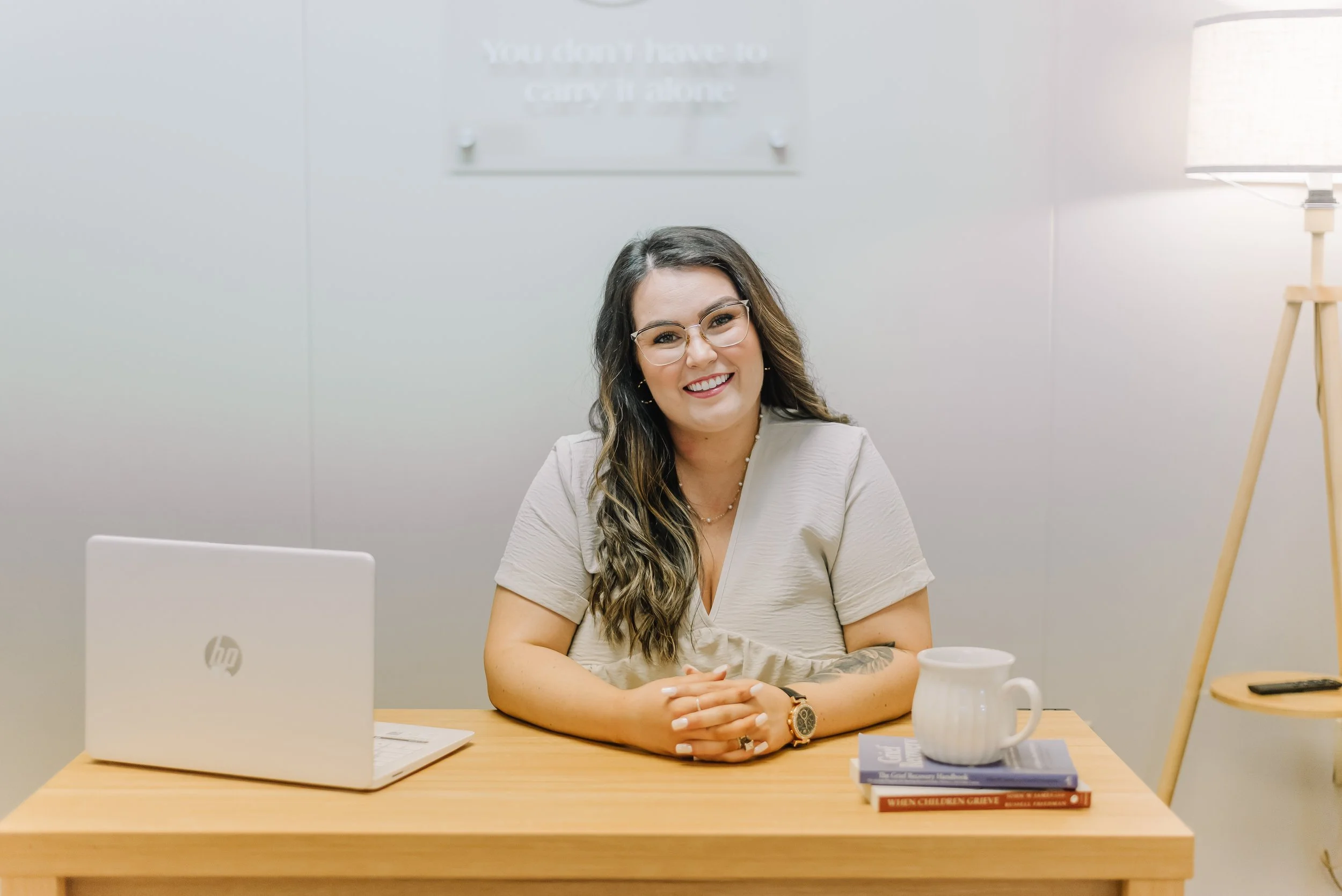 A woman with long wavy hair and glasses smiling at a desk in an office setting, with a laptop, a coffee mug, and books in front of her.