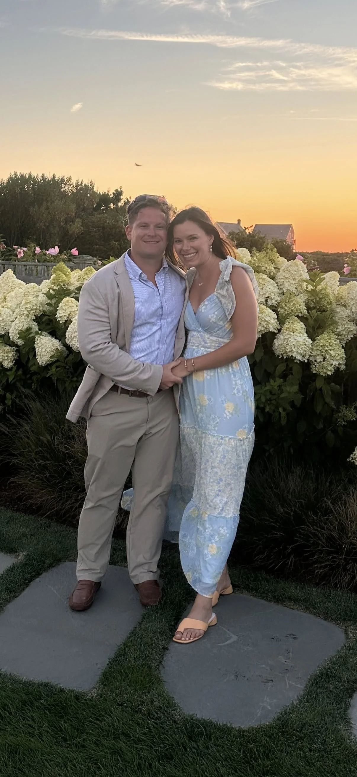 A couple standing outdoors during sunset, in front of white hydrangea bushes. The man is wearing a beige suit with a blue shirt, and the woman is in a long, flowy, light-colored dress with floral patterns. They are holding hands and smiling at the camera.