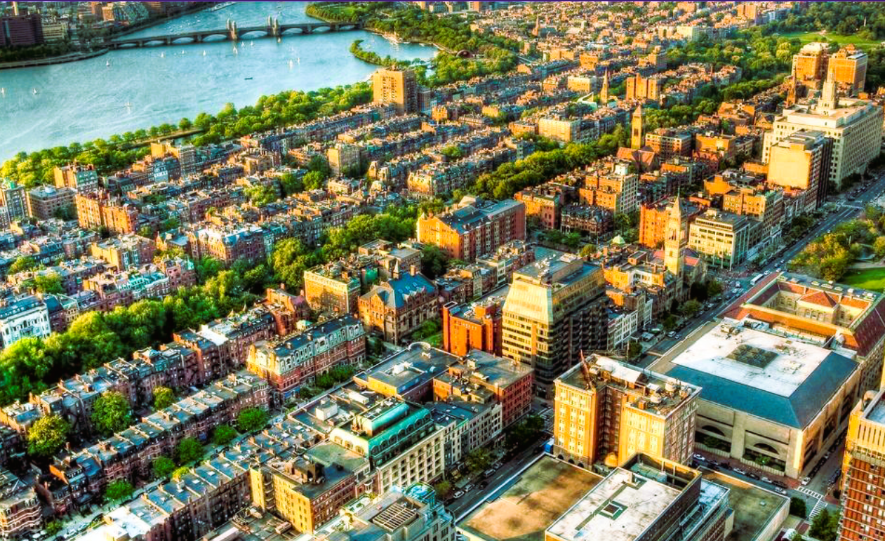 An aerial view of a city with a river, green parks, and a mix of old and modern buildings in the sunlight.