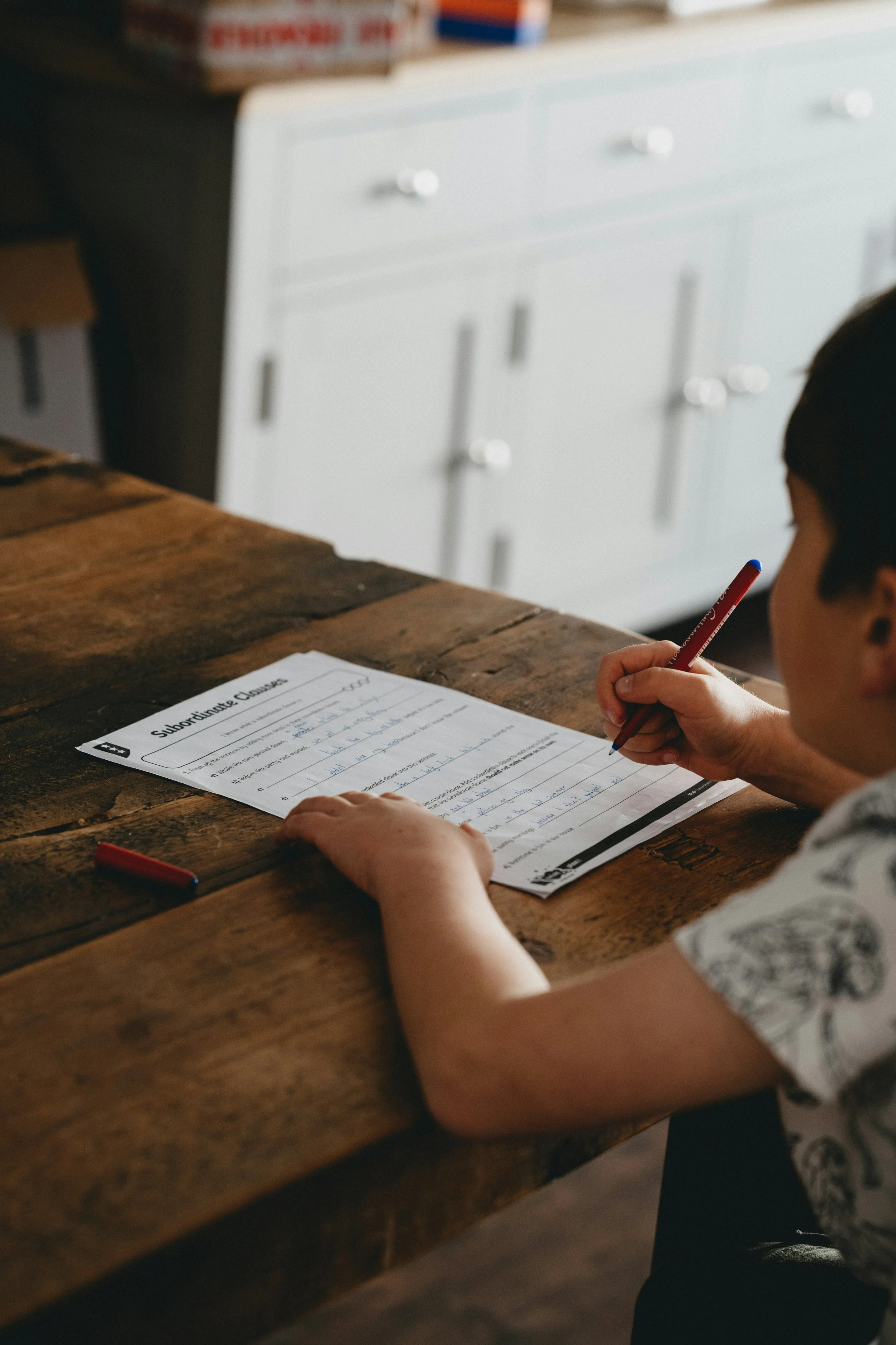 Child writing on a worksheet titled 'Subordinate Clause' at a wooden table.