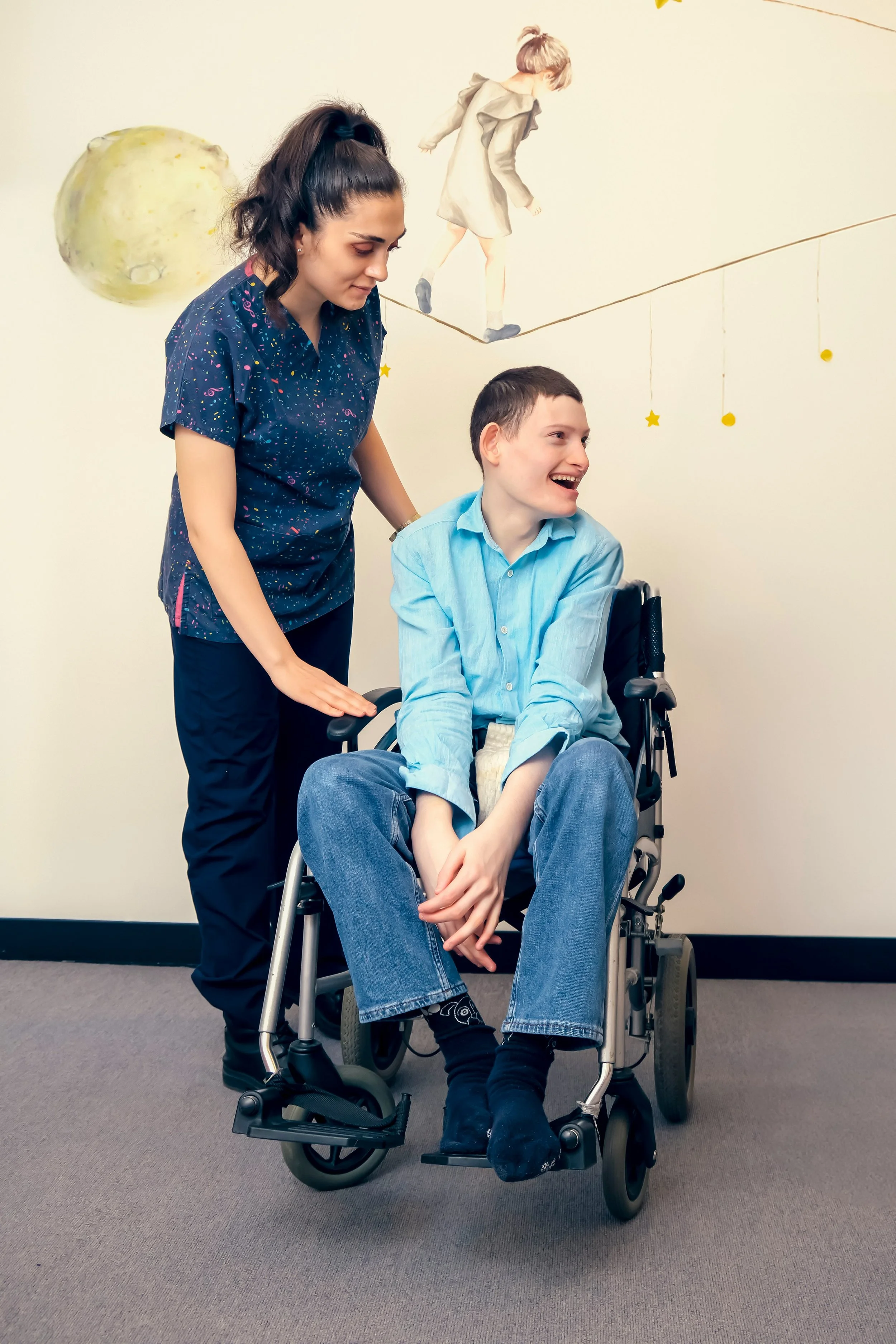 A young woman with long dark hair in a high ponytail stands beside a smiling young man sitting in a wheelchair. The woman has her hand on the wheelchair's armrest as they share a moment of joy. The background features a beige wall with a mural of a girl walking on a line with moon and star decorations.